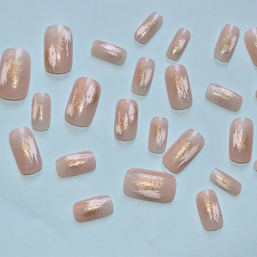 Glossy nude press-on nails featuring white abstract brush strokes and gold foil accents by Soul Struck.