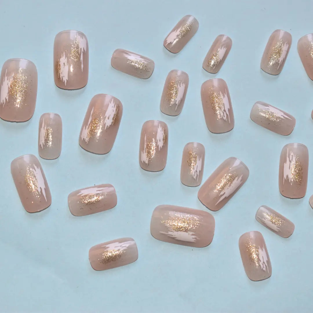 Glossy nude press-on nails featuring white abstract brush strokes and gold foil accents by Soul Struck.